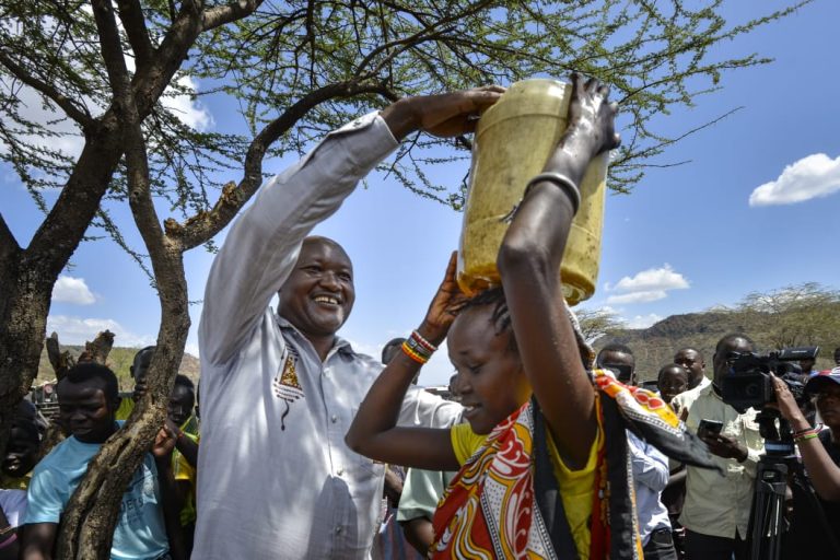 Chief Executive Officer of Mama Doing Good, Dr John Chumo assisting a woman carry a jerrican of water at the Mukur Village waterpoint.