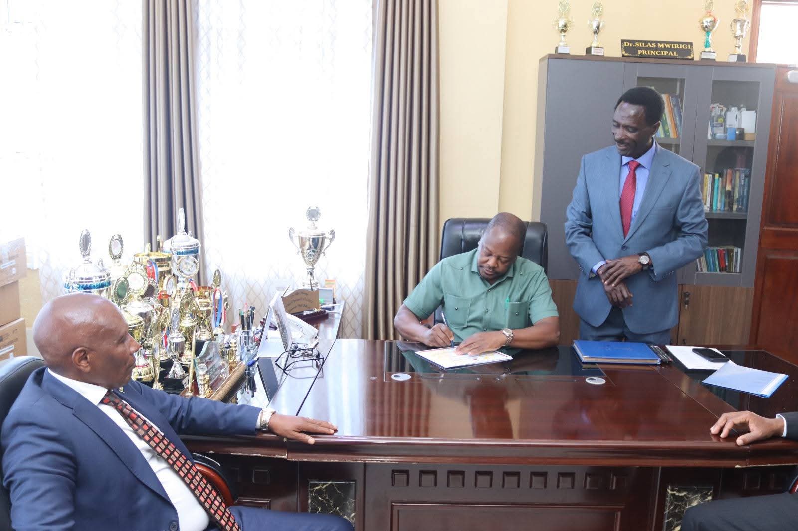 CS Mutahi Kagwe signing Kagumo High School's visitors book as Principal Silas Mwirigi (standing) and KALRO chairman Dr Thuo Mathenge looks on.